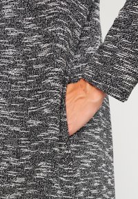 Abrigo de tela texturizada en blanco y negro con un bolsillo integrado visible. El diseño presenta un corte holgado y una suave textura tejida.
