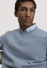 Lichtblauwe gebreide trui met een geribde ronde halslijn, gedragen over een collared wit shirt met subtiele textuur en knoopdetails.