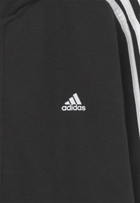 Sweatshirt noir avec logo Adidas blanc ; présente des bandes latérales blanches ; fabriqué en matériau doux ; coupe standard avec un design classique.
