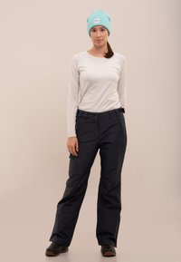 Pantaloni de schi bleumarin cu o croială lejeră, cu buzunare frontale cu fermoar și talie ajustabilă. O bluză albă cu mânecă lungă și o căciulă tricotată teal completează ținuta.