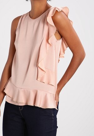 Persoon draagt een mouwloze lichtroze blouse met ruches op de schouders en zoom, gecombineerd met donkerblauwe jeans, staande tegen een effen achtergrond.