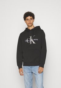 Melnā pārvalka izgatavota no kokvilnas, ar priekšējo kangaroo kabatu un kapuci ar auklu. Iekļauta izteikta baltā Calvin Klein Jeans logo.