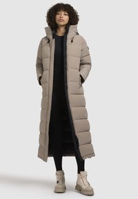 Abrigo largo de plumas beige con capucha, patrón acolchado, cierre de cremallera y bolsillos laterales, usado sobre leggings negros y botas beige de suela gruesa.