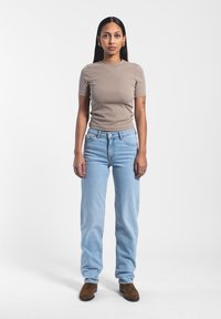 Camiseta ajustada beige, jeans de cintura alta azul claro y botines de ante marrón. Cabello largo y liso; tatuaje notable en el brazo izquierdo.
