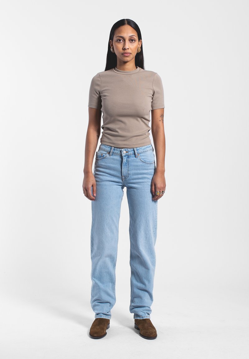 T-shirt beige aderente, jeans a vita alta blu chiaro e stivaletti alla caviglia in suede marroni. Capelli lunghi e lisci; tatuaggio notevole sul braccio sinistro.