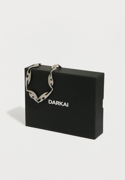 Ogrlica v srebrni barvi z zapletenim dizajnom, ki vključuje motive listov in bleščeče poudarke, prikazana na črni škatli z napisom "DARKAI".