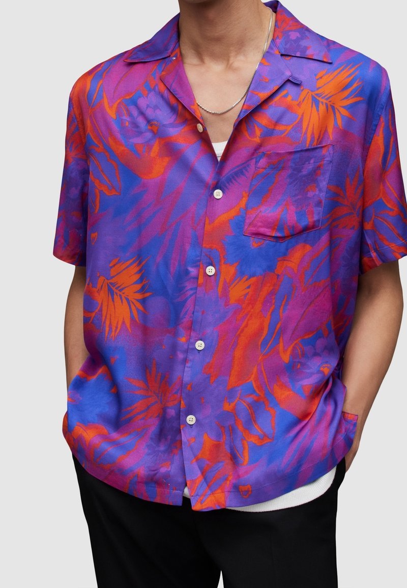 Homme portant une chemise à manches courtes boutonnée à fleurs violettes, orange et bleues, avec une poche poitrine et un collier chaîne argenté.