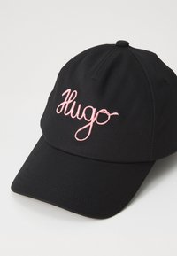 Fekete anyagú sapka íves karimával, amelyen rózsaszín írásos betűkkel hímzett "Hugo" felirat található elöl. Texturált felület varrási részletekkel.
