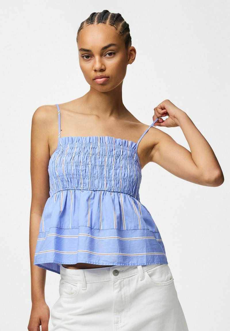Femme avec des tresses cornrow porte un haut bleu smocké sans manches avec de fines bretelles et un pantalon blanc, ajustant une bretelle avec sa main droite.