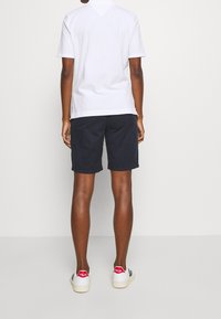 Vit kortärmad t-shirt och marinblå shorts, med en avslappnad passform. Skorna är vita sneakers med svarta ränder och röda hälar.