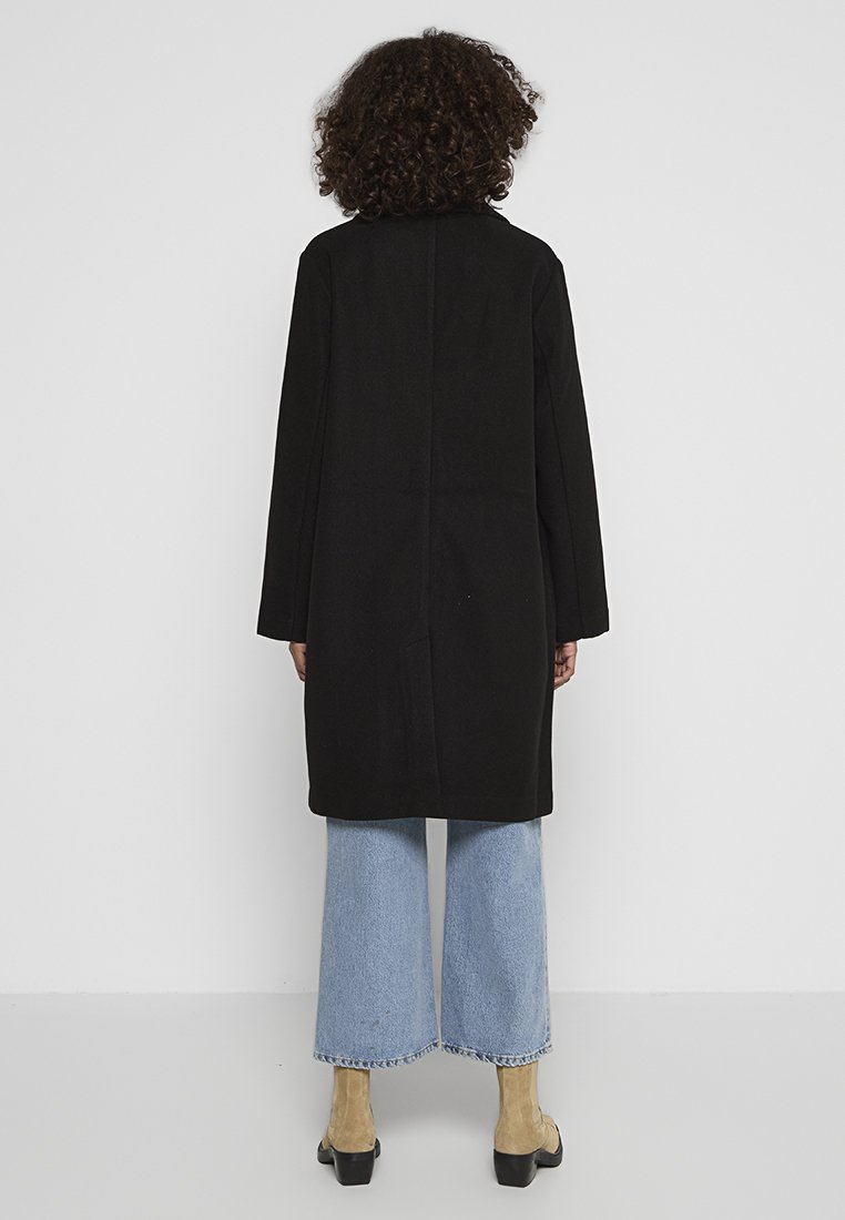 Persoon met krullend haar, gekleed in een zwarte lange jas, lichtblauwe wijde broek en tan kleurige suède hakken, staand met het gezicht naar een witte muur.