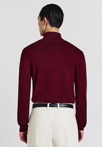 Bordeauxroter Rollkragenpullover mit langen Ärmeln, feiner Struktur, geripptem Kragen, getragen zu beigen Hosen und einem schwarzen Gürtel.