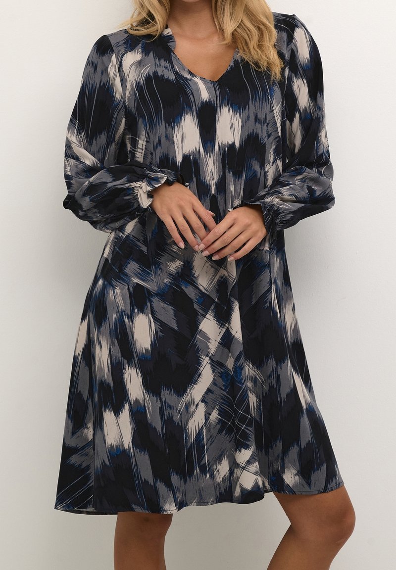 Femme portant une robe à manches longues, longueur genou, avec un motif abstrait noir, gris, blanc et bleu, mains jointes devant.