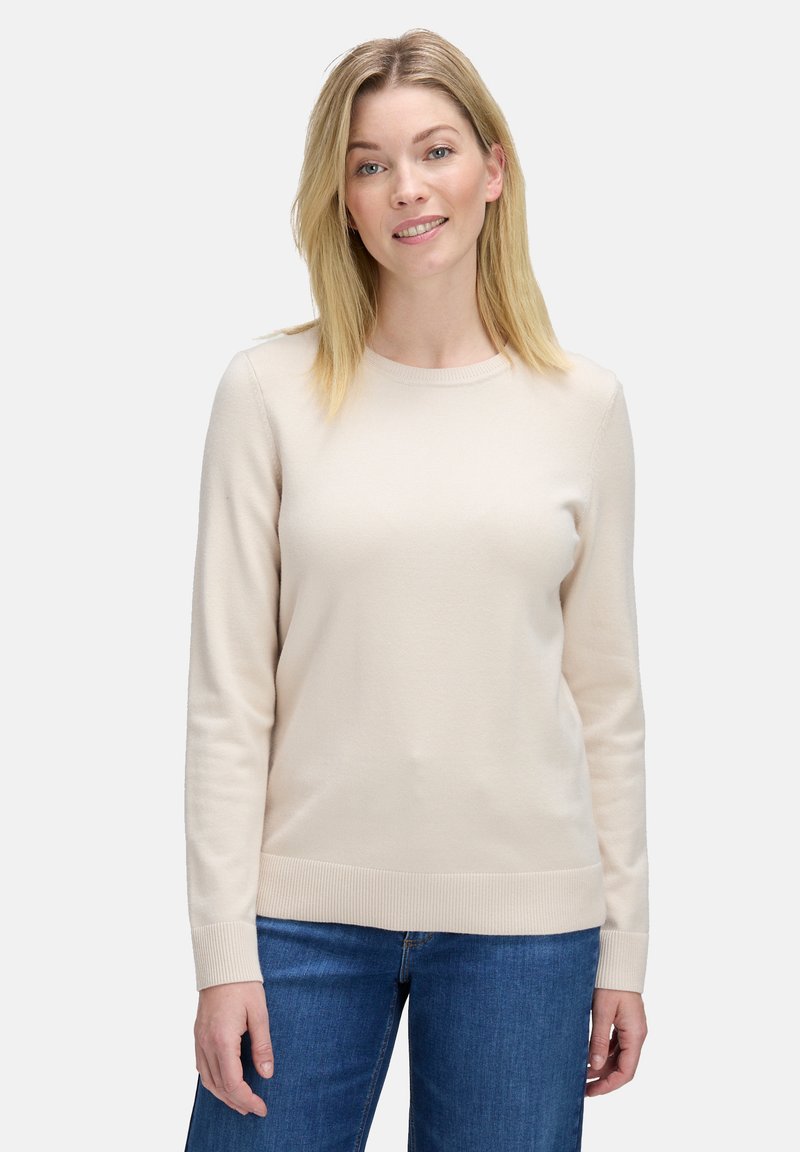 Szőke hajú nő bézs színű, hosszú ujjú pulóvert és kék farmert visel, egy sima világos háttér előtt áll, enyhén mosolyogva.