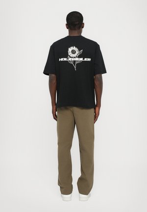 Man facing away wears black T-shirt with white sunflower and "HOLZMEYER" text on back, khaki pants, and white sneakers against plain background.
