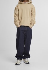 Beige-yliokokoinen huppari, tummansiniset löysät farkut ja vaaleanharmaa-valkoiset lenkkarit. Asussa on etutasku ja huppu.