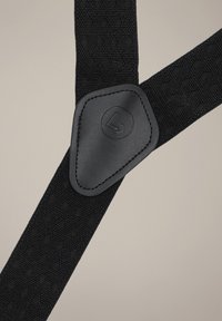 Czarne szelki z tkaniny o teksturowanym wzorze i gładkimi czarnymi skórzanymi akcentami. Posiadają trójkątną skórzaną łatę z logo.