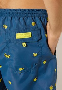 Blauwe zwembroek van lichtgewicht stof, voorzien van gele eendpatronen en een achterzak met een felgeel label.