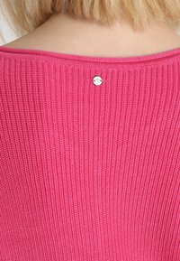 Różowy sweter z dzianiny, o ribbowej teksturze i okrągłym dekolcie. Małe metaliczne logo znajduje się z tyłu.