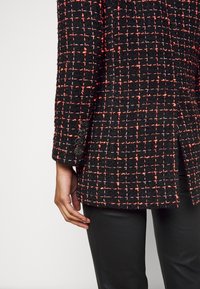Blazer à motif quadrillé noir et rouge avec quatre boutons noirs sur la manche, porté sur un pantalon noir, montrant la main et le bras sur le côté.