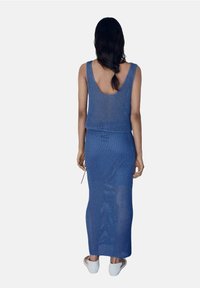 Robe tricotée bleue, sans manches avec un dos en décolleté. Forme longue et ajustée avec un motif texturé. Porta avec des chaussures blanches à enfiler.
