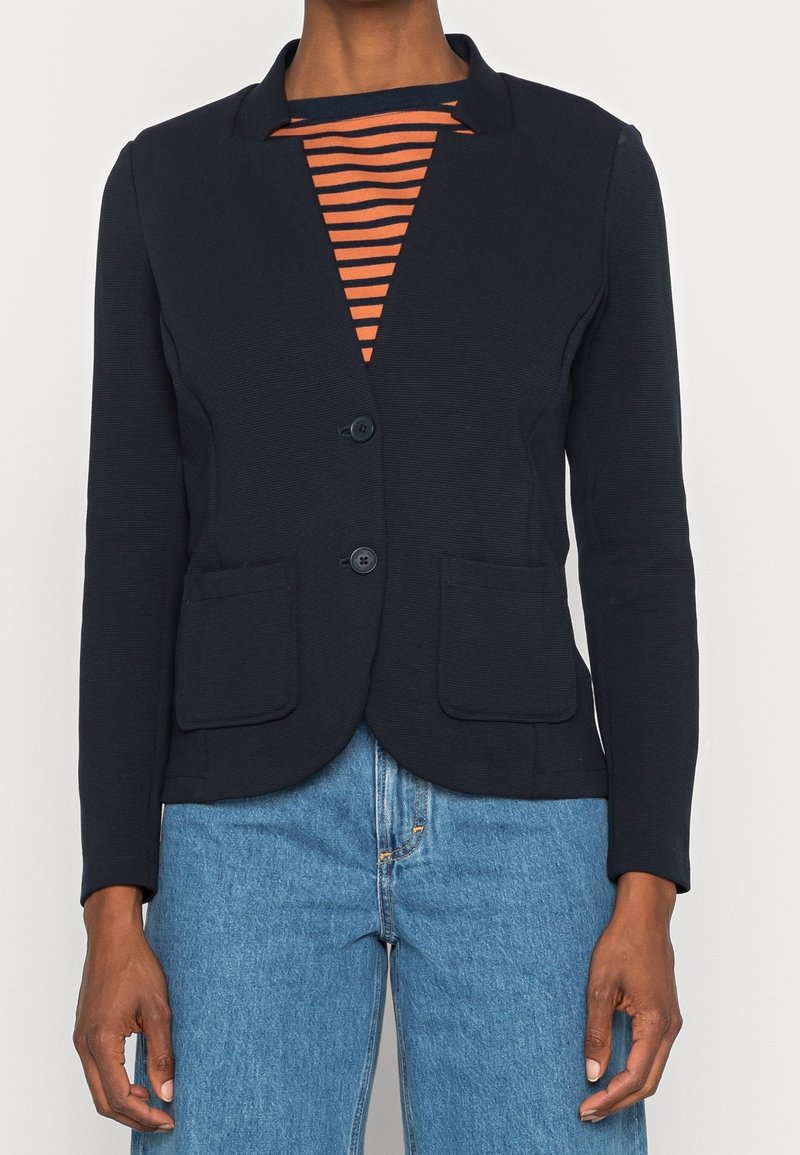 Blazer navy con textura acanalada, dos bolsillos frontales y dos botones. Lleva puesto sobre una camisa rayada naranja y navy, combinada con jeans azules.