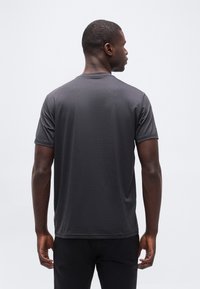 Άνδρας που κοιτάζει προς τα πίσω, φοράει σκούρο γκρι κ οντομάνικο πουκάμισο και μαύρο παντελόνι μπροστά σε μια απλή λευκή επιφάνεια.