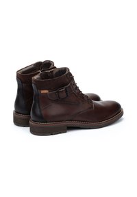Botas de tobillo de cuero marrón con un acabado liso, punta redondeada, acentos oscuros, cordones y un detalle de hebilla. Forro de tela marrón y suela de goma.