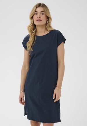 Frau mit langen blonden Haaren, die ein marineblaues, kurzärmliges knielanges Kleid und goldene Creolen vor einem schlichten Hintergrund trägt.