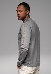 Muž v šedém tričku s dlouhým rukávem, na kterém je nápis na rukávu, stojící na bok s rukama v kapsách na jednobarevném šedém pozadí.