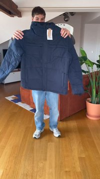 Inimeses hoiab suurt tumesinine talvejacketit, millel on sildid, elutoa keskel, kus on puidust põrand ja potitaim.