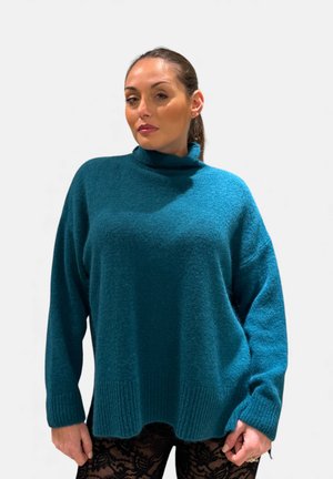 Donna con capelli scuri raccolti in una coda di cavallo, che indossa un maglione dolcevita turchese e pantaloni neri di pizzo, in piedi davanti a uno sfondo bianco uniforme.