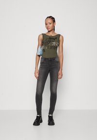 Oliven grønn ribbestrikket tanktop med grafisk print, sammen med mørke grå skinny jeans og svarte kraftige støvler. Hånd holder en liten blå veske.
