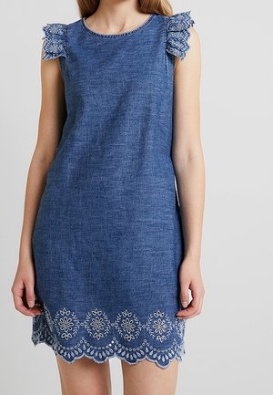 Vestido sin mangas de mezclilla azul con detalles bordados de encaje floral en los hombros y el dobladillo, llevado por una mujer de pie.