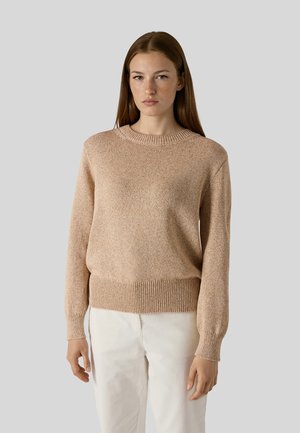 Kvinde med glat brunt hår iført en beige strikket sweater og hvide bukser, stående foran en ensfarvet lys grå baggrund.
