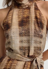 Mouwloze halterjurk met een bruin en beige abstract patroon, voorzien van een hoge halslijn en een strikceintuur in de taille.