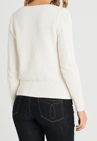 Pull en maille blanche texturée avec ourlet côtelé et détails en tricot torsadé sur les manches. Porté avec un jean foncé ; vue de dos affichée.