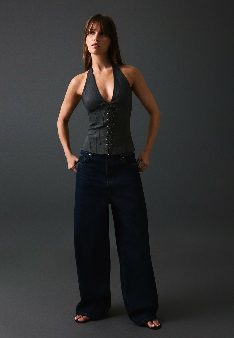 Femme portant un haut corset sans manches gris foncé et un jean large bleu foncé, debout devant un fond sombre uni.