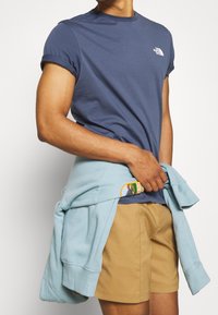 Námořnické bavlněné tričko s krátkým rukávem a malým bílým logem, v kombinaci s světle modrou mikinou uvázanou kolem pasu a béžovými šortkami.