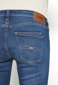 Blue denim jeans feature a brown leather patch labeled "TOMMY JEANS" at the back waist, with contrasting orange stitching and a small logo on the pocket.