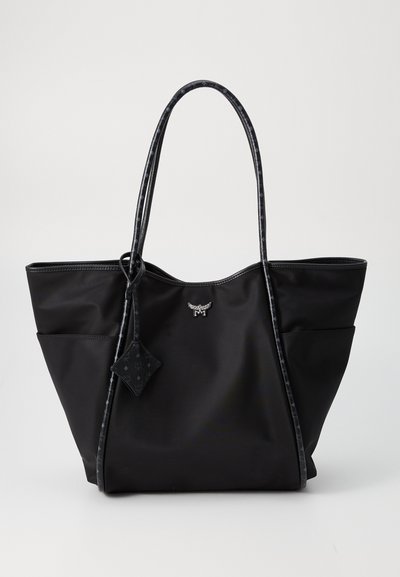 Bolso tote negro con asas largas y decoradas, bolsillos laterales y un pequeño logo metálico en el centro frontal.