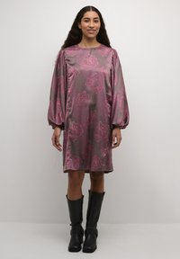 Långärmad klänning i mörkrosasatin med rosa blommönster, med rund halsringning och samlade axlar, tillsammans med knähöga svarta stövlar.