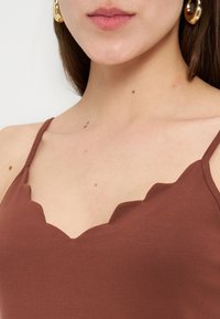 Κοντινό σε λαιμό γυναίκας που φοράει καφέ τοπ με κυματιστή άκρη και χρυσούς κρίκους στα αυτιά, με ορατές ελιές στο στήθος και το λαιμό.