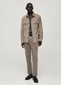Giacca camicia beige con bottoni e due tasche sul petto, abbinata a pantaloni in corduroy beige e scarpe nere, su uno sfondo neutro.