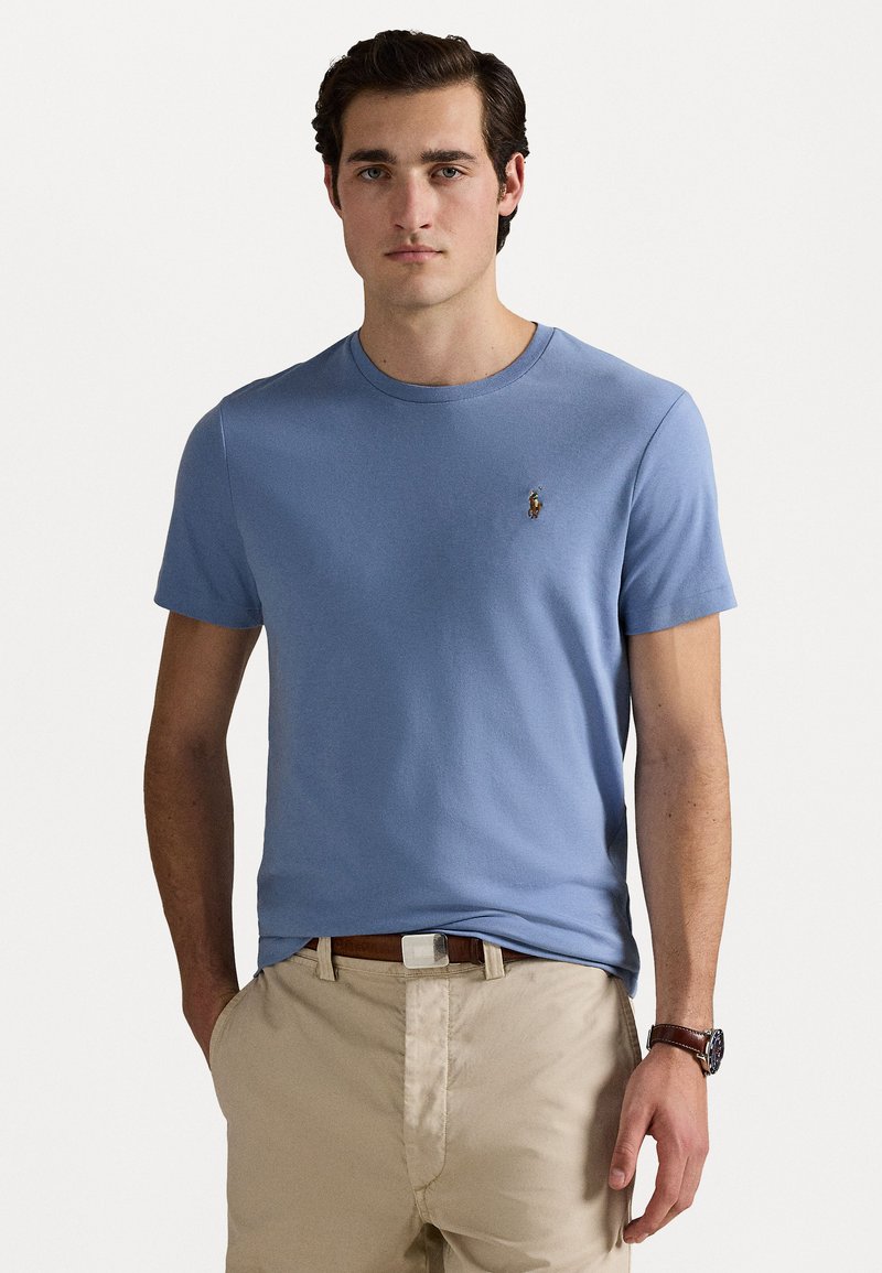 Hele helesinise puuvillane T-särk ümmarguse kaelusega, lühikeste varrukatega ja rinnal väikese tikandiga logoga. Kombineeritud beežide pükstega.