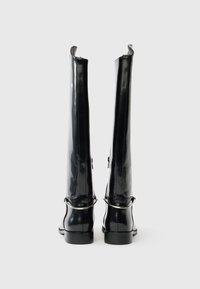 Ζευγάρι ψηλές, γυαλιστερές μαύρες δερμάτινες μπότες με ασημένια διακόσμηση ζώνης στην πίσω πλευρά, όρθιες σε απλό φωτεινό φόντο.