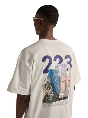 Persoon met kort haar en zonnebril gezien van achteren die een wit T-shirt draagt met nummer 223 en een stads- en berglandschap grafische afbeelding.