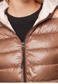 Veste matelassée marron brillante avec une fermeture éclair partiellement ouverte, révélant un pull en maille noir en dessous.