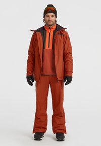 Ski-outfit met een oranje jas, bijpassende broek en zwarte handschoenen. De jas heeft een zak met rits en een kleurgebroken ontwerp.