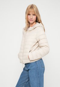 Licht beige pufferjack met een capuchon, ritssluiting en elastische manchetten. Boxy model met horizontale gestikte details; voorzakken zichtbaar.